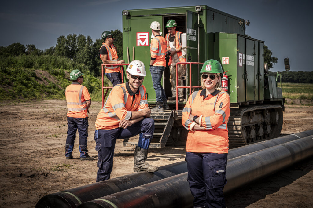 Gruppe von Arbeitern in Sicherheitskleidung auf einer Baustelle mit Maschinen und Rohren.