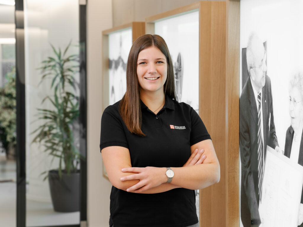 Eine junge Frau mit glattem braunem Haar, die ein schwarzes Poloshirt und eine Uhr trägt, steht lächelnd mit verschränkten Armen in einem modernen Büroflur mit gerahmten Fotos und einer Topfpflanze im Hintergrund.