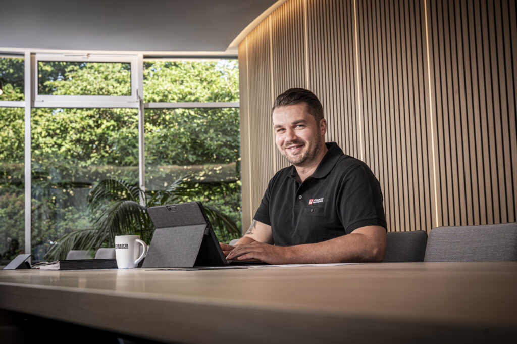 Ein Mann sitzt an einem Konferenztisch und lächelt in die Kamera, während er an einem Laptop arbeitet. Er trägt ein schwarzes Poloshirt mit einem Logo. Große Fenster hinter ihm geben den Blick auf eine grüne, begrünte Außenlandschaft frei. Ein weißer Becher steht auf dem Tisch.