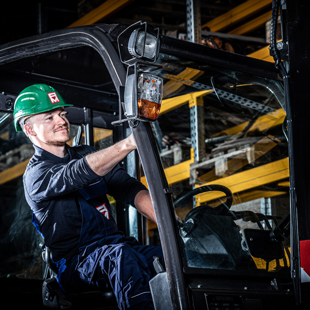 Ein Mann, der einen grünen Schutzhelm und einen blauen Arbeitsoverall trägt, bedient schwere Maschinen in einem Lagerhaus mit gelben Regalen und Holzpaletten im Hintergrund.