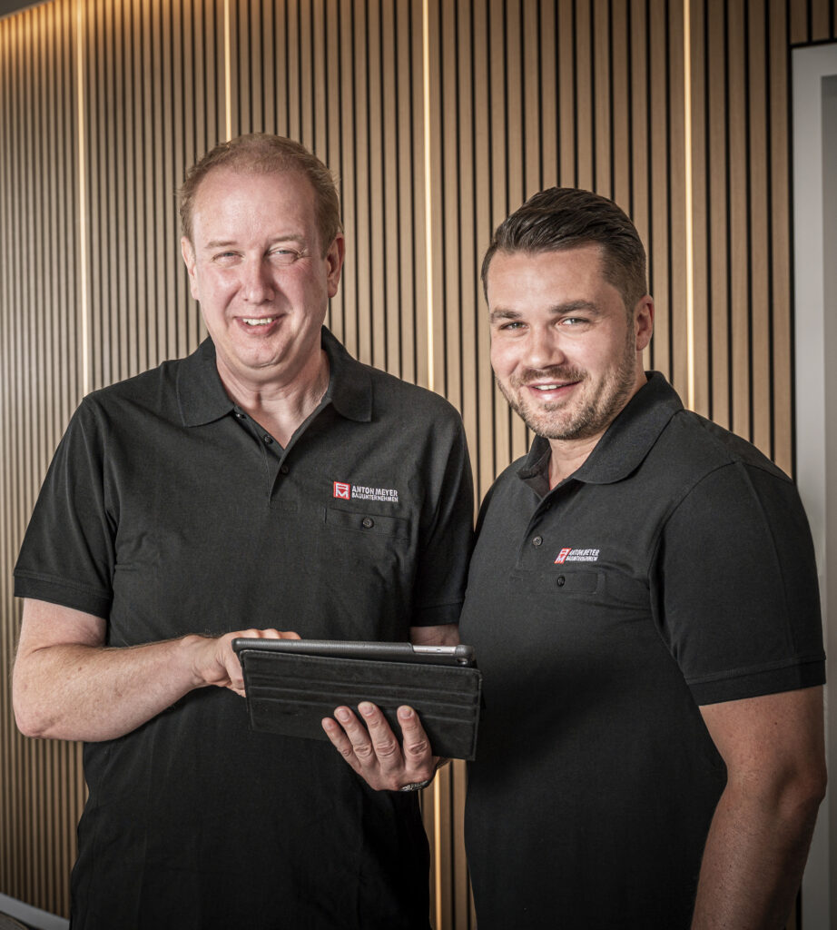 Zwei Männer in identischen schwarzen Poloshirts mit Anton-Meyer-Logos, von denen einer ein Tablet in der Hand hält, stehen vor einer Wand aus Holzlatten.